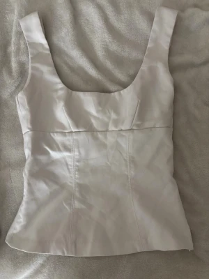 Vit topp  med breda axelband - Snygg vit topp med breda axelband och markerade sömmar som ger en figurnära passform. Har en dragkedja på sidan. Materialet är lite som jeansmaterial. Säljer då den är för liten för mig💗