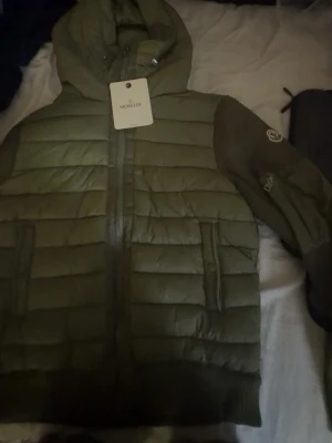 Olivgrön dunjacka  - Snygg olivgrön dunjacka med huva och dragkedja framtill. Jackan har två fickor med lock och ribbade muddar vid ärmslut och nederkant. Klassisk logga på ärmen och quiltad design för extra värme.