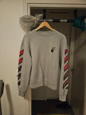 Grå sweatshirt med röda detaljer - Säljer en grå sweatshirt med tryckta röda och svarta grafiska detaljer på ärmarna och en stor grafisk design på ryggen. Tröjan har rund hals och lång ärm, perfekt för en streetwear-look.