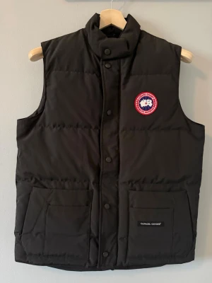 Svart dunväst från Canada Goose - Stilren svart dunväst från Canada Goose med hög krage och tryckknappar framtill. Västen har två stora fickor och den klassiska Canada Goose-loggan på bröstet. Perfekt för lager-på-lager och håller dig varm under kyliga dagar.