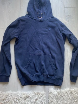 Marinblå hoodie från U.S. Polo Assn - Säljer en marinblå hoodie från U.S. Polo Assn med klassisk känguruficka och broderad logga på bröstet. Hoodien har långa ärmar, ribbade muddar och är tillverkad i mjuk bomull. Perfekt för en chill och avslappnad stil.