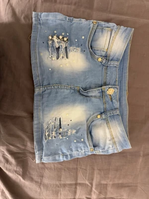 Ljusa jeansshorts med Union Jack-detalj - Säljer ett par ljusblå jeansshorts med slitningar, pärldetaljer och Union Jack-flagga på bakfickan. Shortsen har guldiga knappar och klassisk femficksdesign. Perfekta för dig som vill sticka ut i sommar!