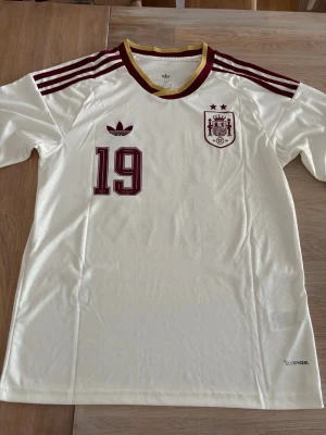 Spaniens matchtröja Adidas nr 19 s - Säljer en vit Spanien matchtröja från Adidas med vinröda detaljer, nummer 19 och Lamine Yamal på ryggen. Tröjan har korta ärmar, rund hals med gul kant och Climacool-material för bra ventilation. Perfekt för fotbollsfans! Längden: 68 cm och bredden 46!