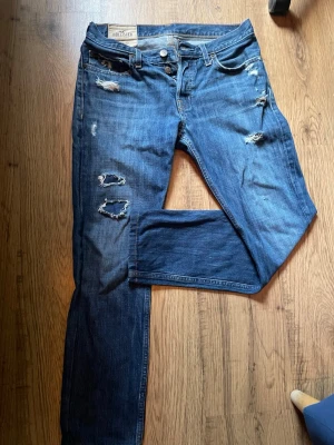 Blå slitna jeans från Hollister - Säljer ett par blå jeans från Hollister med skinny fit och flera slitningar och hål på benen för en trendig, avslappnad look. Klassisk femficksmodell med knappgylf och låg midja. Perfekta för dig som gillar streetstyle och casual vibes.