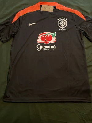 Brasilien träningsset  - Säljer ett svart träningsset från Nike med Brasilien-logga och Guaraná Antarctica-tryck. Tröjan har röda axelpartier och shorts med röda detaljer på sidorna. Perfekt för fotbollsträning eller gymmet. Materialet är lätt och andas.