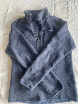 Mörkblå fleece från Patagonia - Mysig mörkblå fleece med halv dragkedja från Patagonia. Tröjan har hög krage, bröstficka med dragkedja och klassisk logga på bröstet. Perfekt för lager-på-lager och outdoor-looks. Storlek S