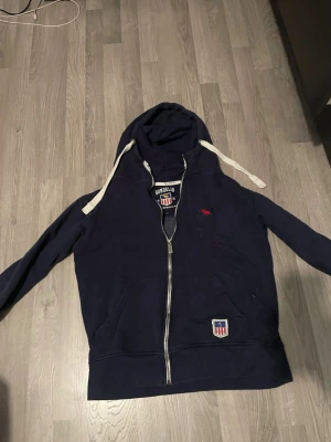 Mörkblå hoodie från Bondelid - Mörkblå hoodie från Bondelid med dragkedja, vita snören i huvan och två framfickor. Märkets logga och emblem finns på bröstet och nedtill. Hoodien har en klassisk passform och är tillverkad i mjukt material, perfekt för en avslappnad stil. OBS storlek M men sitter som s