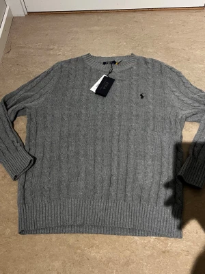 Grå kabelstickad tröja Polo Ralph Lauren - Kabelstickad grå tröja från Polo Ralph Lauren med rund halsringning och svart broderad logga på bröstet. Tröjan har långa ärmar och ribbade muddar vid ärmslut och nederkant. Perfekt för lager på lager och klassisk stil.