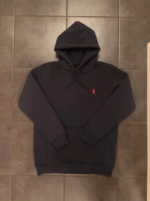 Mörkblå hoodie från Polo Ralph Lauren - Snygg mörkblå hoodie från Polo Ralph Lauren med klassisk röd logga broderad på bröstet. Aldrig använd, är helt ny. Hoodien har huva med dragsko, magficka och ribbade muddar vid ärmar och nederkant. Perfekt för en chill och stilren look.