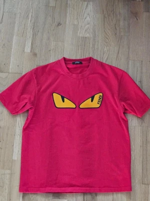 Röd Fendi t-shirt med ögontryck - Cool röd t-shirt från Fendi med ikoniskt ögontryck i gult och svart på bröstet. Klassisk passform med rund hals och korta ärmar. Perfekt statement-plagg för dig som vill sticka ut. Tillverkad i mjuk bomull.