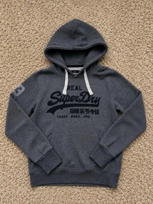 Mörkgrå Superdry hoodie med tryck - Snygg mörkgrå hoodie från Superdry med svart logotyptryck och japanska tecken på bröstet. Klassisk känguruficka, huva med vita snören och ribbade muddar. Perfekt för en avslappnad streetstyle-look.