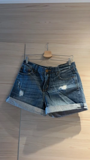 Blå denimshorts med upprullad kant - Blå jeansshorts i klassisk denim med upprullade benslut och slitna detaljer framtill. Shortsen har fem fickor och knappgylf. 100% bomull