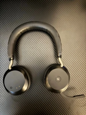 Svarta over-ear hörlurar med mikrofon (Jabra Evolve) - Stilrena svarta over-ear hörlurar med rund design och mjuka öronkuddar. Utrustade med en mikrofonarm och vadderad bygel i konstläder för extra komfort. Perfekta för gaming, samtal eller musiklyssning. Trådlös design och diskreta knappar på sidan.