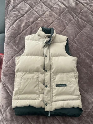 Vintage Canada Goose dunväst – Beige – Äkta – Mycket bra skick - Säljer en snygg vintage Canada Goose-väst i beige med svart insida och den äldre Canada Goose Expedition Clothing Outfitters-patchen.  Västen är äkta och i väldigt bra skick. Klassisk äldre modell med stora framfickor, tryckknappar och rejäl vaddering.  📏 Storlek XS, men sitter mer som en S.  En ovanligare vintage-modell som inte längre säljs i denna design. Motsvarande Canada Goose-västar kostar idag ca 7 000–8 000 kr nya.