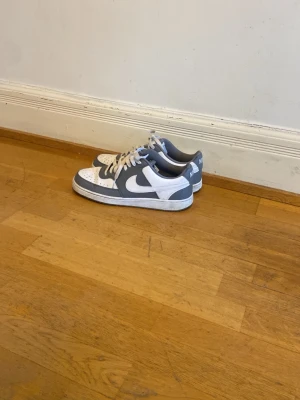 Nike Dunk Low blå och vita sneakers - Snygga Nike Dunk Low sneakers i grått och vitt med klassisk swoosh på sidan. Skorna har perforerad tåbox, vita skosnören och en låg siluett som ger en clean och sportig vibe. Perfekta för dig som vill ha en stilren och trendig look. Använt 10 gånger cirka orginal pris 1100