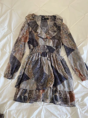 Mönstrad kortklänning från Zara - Kortklänning från Zara med patchwork-inspirerat mönster i blå, beige, brun och grå toner. Klänningen har volangdetaljer vid halsringningen, långärmade ärmar med resår vid handlederna och smock i midjan för en snygg passform. Tunt, lätt material som ger en luftig känsla.