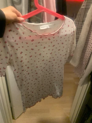 Vit blommig topp från Name It - Söt vit topp från Name It med små rosa blommor och vågig kant i rosa vid hals och ärmslut. Toppen är ribbad och har korta ärmar, perfekt för en avslappnad och trendig look. Passar dig som gillar gulliga detaljer och mjuka material.