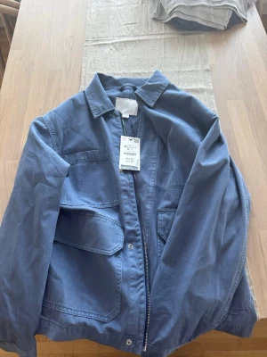 Blå overshirt med dragkedja - Snygg blå overshirt i bomull med dragkedja framtill och två stora fickor med lock. Klassisk krage och avslappnad passform, perfekt att slänga på sig över en t-shirt. Enkel och stilren design som funkar till många outfits.