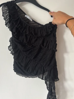 Svart oneshoulder topp med volanger - Säljer en svart oneshoulder topp  från Zara! Det står storlek S men passar xs ockås💗