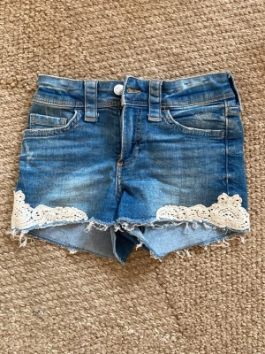 Blå jeansshorts med spetsdetaljer - Snygga blå jeansshorts med rå kant och vita spetsdetaljer längst ner på benen. Klassisk femficksmodell med bälteshällor och knappgylf. Perfekta för varma dagar och ger en avslappnad men söt look. Från H&M