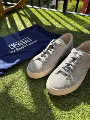 Gråa sneakers från Polo Ralph Lauren - Snygga gråa sneakers i mocka från Polo Ralph Lauren. Skorna har klassisk låg profil, vita sulor och matchande grå skosnören. Perfekta för en clean och stilren look. Kommer med original dustbag. Använda 1 gång då jag kom på att dom var för stora så inga fläckar elr nått sånt. 