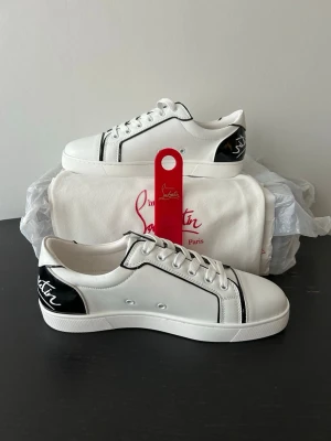 Vita Louboutin sneakers med svart detalj - Snygga vita sneakers från Christian Louboutin med svarta detaljer och logga på hälen. Skorna har klassisk låg profil, vita snören och den ikoniska röda sulan. Tillverkade i slätt läder med svarta kontrastlinjer för en clean och modern look. Allt OG medföljer förutom OG box