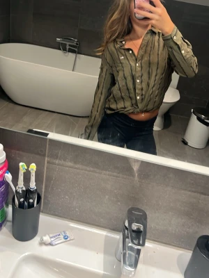 Randig skjorta från Tommy Jeans - Säljer en grön skjorta från Tommy Jeans med svarta vertikala ränder och vita knappar. Skjortan har en bröstficka med liten logga och klassisk krage. Perfekt för dig som gillar en avslappnad men ändå stilren look.
