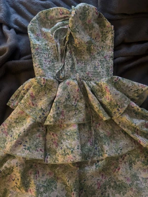 Blommig volangklänning med snörning - Supersöt blommig klänning med volanger och smockad överdel. Klänningen har tunna axelband med snörning och omlott-detalj vid bysten. Färgerna går i ljusgrönt, vitt och inslag av rosa och lila. Perfekt för sommarens alla tillfällen!