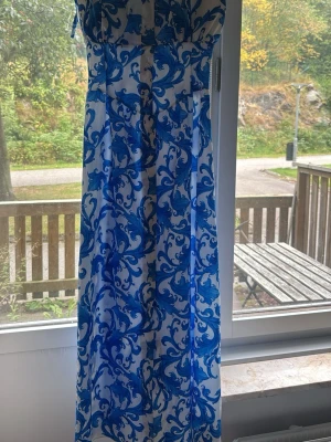 Vit långklänning med blått mönster - Säljer en vit långklänning med tunna axelband och ett iögonfallande blått mönster. Klänningen har knappar framtill och ett flowy tyg som ger en luftig känsla. Perfekt för varma dagar och somriga tillfällen.