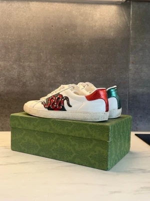 Skor Gucci Ace Snake i toppenskick - Trendiga Gucci sneakers med broderad röd och svart orm på sidan. Hälen har metallicdetaljer i rött och grönt för extra edge. Skorna har klassisk låg profil och vita snören. Perfekta för dig som vill sticka ut med coola detaljer och unik design.