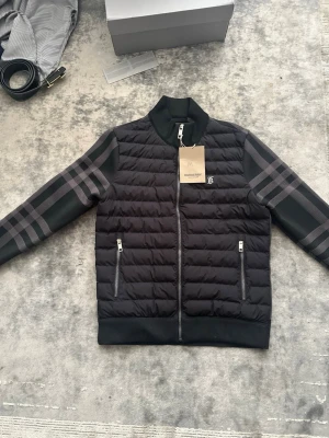 Svart pufferjacka från Burberry - Säljer en svart pufferjacka från Burberry med klassiskt rutmönster på ärmarna och diskret logga på bröstet. Jackan har ribbade muddar, dragkedja framtill och två fickor med dragkedja. Materialet är quiltat och ger en modern look.