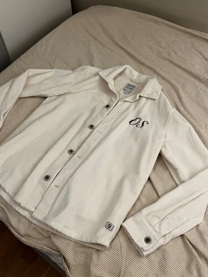Vit manchester overshirt från Only & Sons - Vit overshirt i manchester med svarta broderade bokstäver på bröstet. Skjortan har klassisk krage, knappar framtill och långa ärmar med knapp vid manschetten. Perfekt för dig som gillar stilrena detaljer och mjukt material.