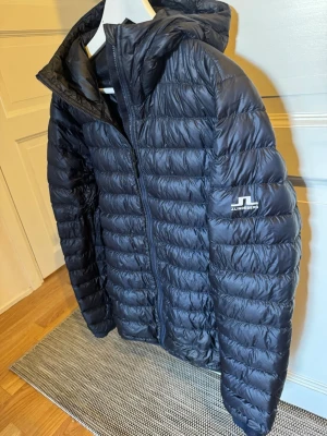 Svart dunjacka från J.Lindeberg - Snygg svart dunjacka från J.Lindeberg med huva och dragkedja framtill. Jackan har quiltad design och logga på ärmen. Perfekt för kalla vinterdagar och riktigt lätt att bära. Passar dig som vill ha både stil och funktion.