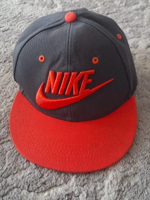 Nike snapback keps grå och röd - Snygg snapback keps från Nike i grått med röd skärm och röda detaljer. Stor Nike-logga broderad framtill. Justerbar baktill med plastspänne. Tillverkad i bomullstyg, perfekt för en sportig och avslappnad stil.