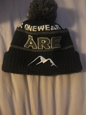 Åre mössa - Säljer en svart stickad mössa med grå och vita detaljer, stor tofs på toppen och texten 'ÅRE' samt 'ONEWEAR' runt om. Mössan har ett broderat bergmotiv framtill och bred uppvikt kant. Perfekt för kalla dagar i fjällen eller stan.