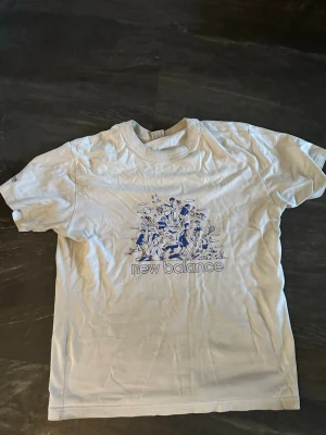 Vit t-shirt från New Balance - Vit t-shirt från New Balance med ett stort blått tryck framtill som visar olika sportaktiviteter och texten 'new balance' under. Klassisk passform och rund hals. Perfekt för dig som gillar sportig stil och streetwear.