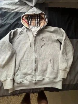 Burberry hoodie  - Säljer en grå zip-hoodie med klassiskt rutigt foder i huvan. Hoodien har dragkedja framtill, två stora fickor och ribbade muddar vid ärmar och nederkant. Perfekt för chill dagar och enkel att matcha med det mesta.