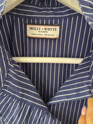 Blåvit randig blus Holly & Whyte - Snygg blå blus med vita vertikala ränder från Holly & Whyte. Blusen är ärmlös med volangdetaljer vid axlarna och har en dekorativ krage. Perfekt för en trendig och fräsch look. Tillverkad i ett lätt och luftigt material.