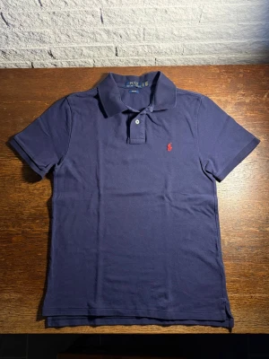 Ralph Lauren Piké - Klassisk marinblå pikétröja från Ralph Lauren med röd broderad logga på bröstet. Hör av dig vid eventuella frågor!