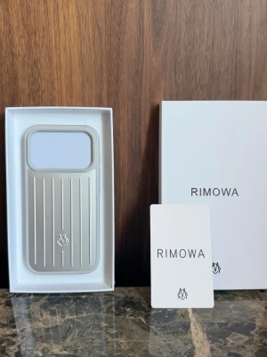 Silverfärgat mobilskal från Rimowa 17 pro - Snyggt och stilrent mobilskal från Rimowa i silverfärgad aluminium med klassiska räfflor och logotyp på baksidan. Skalet har en rektangulär form med stor öppning för kameran och är designat för att ge en exklusiv känsla. Perfekt för dig som gillar minimalistisk och modern design.