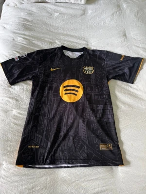 Svart Barcelona matchtröja Nike Lamine Yamal  - Snygg svart Barcelona matchtröja med gula detaljer och tryck, inklusive Spotify-logga på bröstet och UNHCR-logga på ryggen. Nummer 19 och namnet Lamine Yamal på ryggen. Korta ärmar och stadsmotiv i bakgrunden. Perfekt för fotbollsfans! Pris kan diskuteras