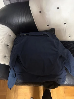 Mörkblå vindjacka med rutigt foder - Snygg mörkblå vindjacka med klassisk krage och rutigt foder på insidan. Jackan har knappar vid ärmsluten och dragkedja framtill. Perfekt för dig som vill ha en stilren och enkel jacka till vardags.