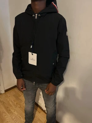 Svart Moncler vindjacka med huva - Snygg svart vindjacka från Moncler med huva och dragkedja framtill. Jackan har två fickor med knappar och en vit-röd rand på huvan. Perfekt för blåsiga dagar och enkel att matcha med jeans eller sneakers. Jackan är i storlek 1 som motsvarar storleken S!!😀