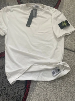 Vit t-shirt från Stone Island - Vit t-shirt från Stone Island med klassisk rund hals och korta ärmar. På vänster ärm sitter den ikoniska svarta patchen med gult och grönt Stone Island-märke. Tillverkad i mjuk bomull för en clean och stilren look.