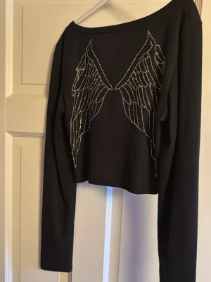 Svart croppad topp med änglavingar - Snygg svart croppad långärmad topp med två knappar framtill och fina änglavingar av glittriga stenar på ryggen. Perfekt för dig som vill sticka ut med en unik detalj. Toppen har en lätt ribbad struktur och sitter normal på kroppen.