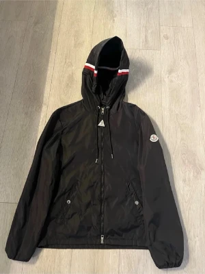Svart Moncler vindjacka med huva - Svart vindjacka från Moncler med huva och dragsko. Jackan har dragkedja framtill, två fickor med knappar och Moncler-logga på ärmen. Huvan har en vit och röd rand som detalj. Perfekt för blåsiga dagar och snygg streetstil.