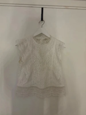 Vit spetsblus med volangdetaljer - Superfin vit blus i spets med volangdetaljer vid ärmarna och nederkanten. Blusen är ärmlös och har en rund halsringning. Perfekt att styla med jeans eller kjol för en fräsch look.