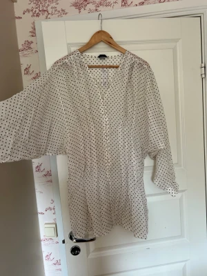 Vit prickig blus med knappar- oanvänd - Säljer en luftig vit blus med bruna prickar och knäppning framtill. Blusen har v-ringning och vida långärmar. Materialet känns tunt och flowy, perfekt för en chill och trendig look. Passar till både jeans och kjol. Oanvänd- orginalpris 399