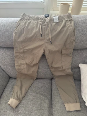 Beiga cargobyxor från H&M - Snygga beiga cargobyxor från H&M med resår i midjan och dragsko. Byxorna har stora sidofickor och muddar vid bensluten. Tillverkade i en lätt och bekväm bomullsblandning, perfekta för en avslappnad stil.