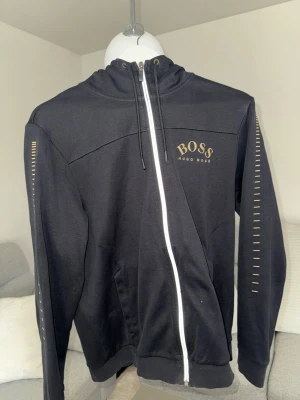 Svart hoodie från BOSS med dragkedja - Snygg svart hoodie från BOSS med vit dragkedja och guldfärgad logga på bröstet. Hoodien har huva med dragsko och diskreta detaljer längs ärmarna. Perfekt för en stilren och avslappnad look.